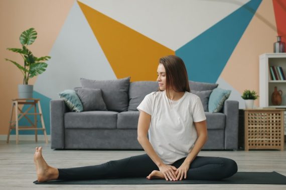 Attractive girl in leggings and T-shirt is doing stretching exercises on yoga mat relaxing on floor at home busy with practise. Youth, lifestyle and health concept.