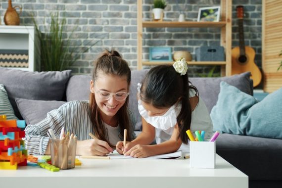 Happy little girl is drawing at home with caring mother using pencils having fun in modern apartment. Creative leisure activity and childhood concept.